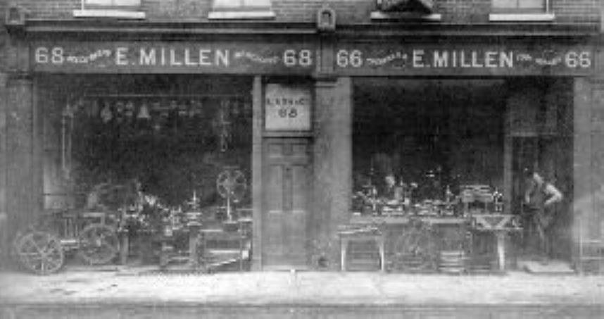 Black and white photograph of the original E. Millen machine tool shop at numbers 66 and 68, with lathes and machinery in both windows and a man standing in the right-hand doorway.